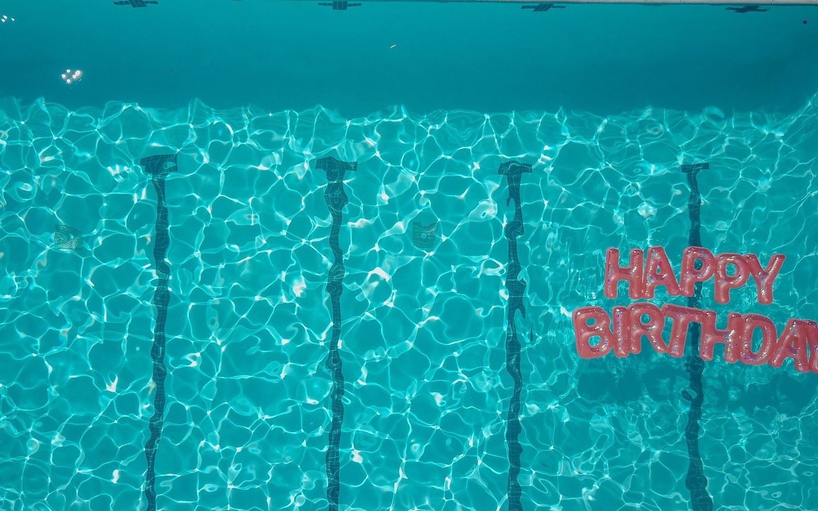 Red "happy birthday" balloon letters float on rippling turquoise pool water with lane markers